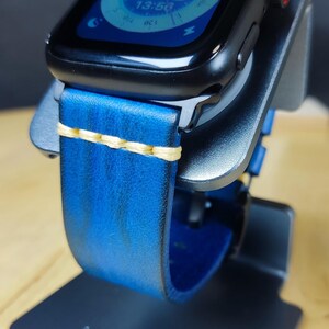 Puede incluir: Un Apple Watch negro con una correa de cuero azul y un detalle de costura blanca. El reloj está en un soporte de metal gris con la palabra "Lemnos" impresa en él. La esfera del reloj muestra un reloj digital con la hora 13:58.