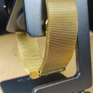 May include: A gold metal watch band with a mesh design. The band is on a gray metal watch stand.
