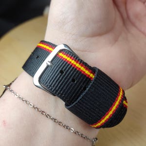 May include: Black watch strap with red and yellow stripes. The strap is made of a woven material and features a silver-colored buckle. A silver chain bracelet is also visible.