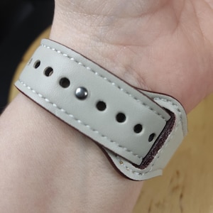 May include: A light gray leather watch band with a burgundy underside and white stitching. The band has multiple holes for adjustment and a silver-colored metal clasp. The watch band is shown on a wrist.