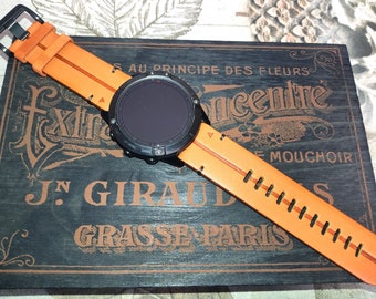 May include: A black smartwatch with an orange leather band. The watch face is black and the band has orange stitching. The watch is on a wooden surface with French text.