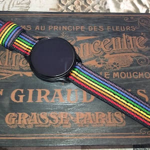 May include: A black smartwatch with a rainbow-striped nylon band. The band features horizontal stripes of red, orange, yellow, green, blue, and purple. The watch is displayed on a dark wooden surface with vintage text.