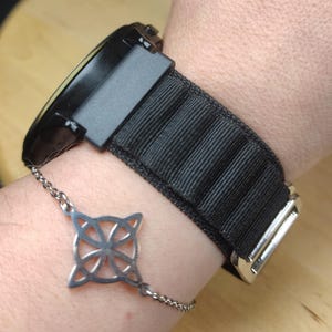 Puede incluir: Un reloj negro con una correa de nailon negra y una hebilla plateada. El reloj está en la muñeca de una persona y hay un brazalete de cadena plateada con un diseño de nudo celta en la otra muñeca.
