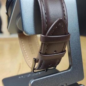 May include: A brown leather watch band with white stitching is shown on a silver watch stand. The band has the words "Genuine Leather" printed on the inside.