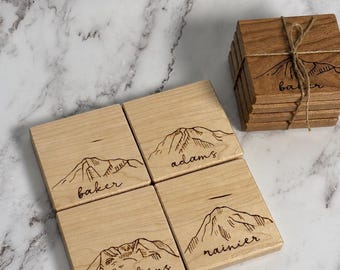 May include: A set of square light-colored wooden coasters featuring laser-engraved mountain illustrations and the names baker, adams, st. helens, and rainier. One stack of coasters is tied together with thin brown twine.