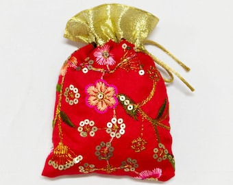 May include: A red drawstring pouch with gold accents. The pouch is adorned with embroidered floral patterns in pink, gold, and green, and embellished with sequins. The top of the pouch is gold and the drawstring is a matching gold color.