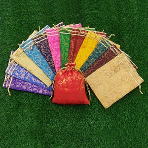 May include: A collection of colorful drawstring gift bags in various shades, including red, gold, blue, and purple. Each bag features a gold floral pattern and a gold drawstring closure. The bags are arranged on a green surface.