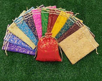Könnte beinhalten: Eine Sammlung farbenfroher Geschenkbeutel mit Kordelzug in verschiedenen Farbtönen, darunter Rot, Gold, Blau und Lila. Jeder Beutel hat ein goldenes Blumenmuster und einen goldenen Kordelverschluss. Die Beutel liegen auf einer grünen Oberfläche.