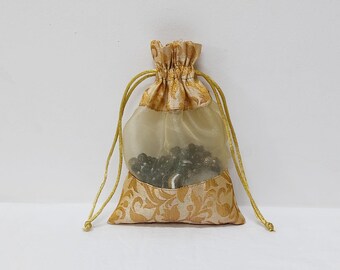 Könnte beinhalten: Eine kleine, goldene Kordeltasche aus durchsichtigem Stoff mit floralem Muster. Die Tasche ist mit schwarzen Perlen gefüllt und hat ein weißes Satin-Futter.