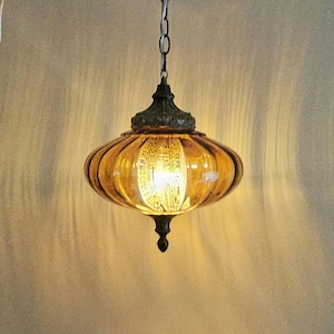 Può includere: Una lampada a sospensione con un paralume in vetro color ambra a coste, dalla forma arrotondata e appiattita. L'apparecchio presenta un cappuccio in metallo scuro e decorato sulla parte superiore e un terminale abbinato alla base, sospeso da una catena.