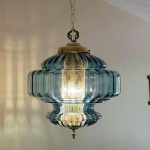 May include: A hanging pendant light fixture featuring a ribbed, translucent blue glass shade with a tiered, rounded silhouette. The lamp is suspended by a gold-toned metal chain and includes a central brass-colored cap and a small decorative finial.