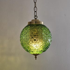May include: A green glass pendant light with a spherical shade featuring an embossed floral pattern. The light fixture is suspended by a brass chain and has brass accents. The light is illuminated, casting a warm glow.