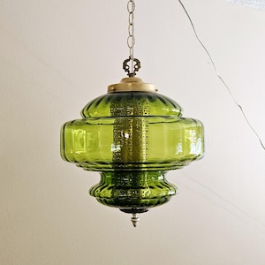 May include: A vintage green glass pendant light fixture. The light has a brass-colored top and chain, with a decorative finial at the bottom. The glass shade is a vibrant green color and has a unique, layered design. The fixture hangs from a chain.