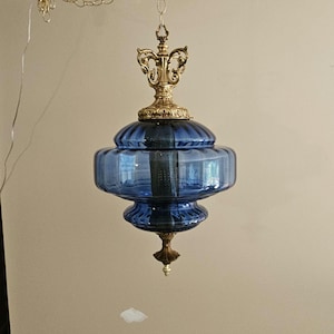 May include: A hanging pendant lamp featuring a translucent blue glass shade with a ribbed, tiered design. The fixture includes ornate, metallic gold-toned hardware at the top and bottom, suspended by a matching gold-toned metal chain.