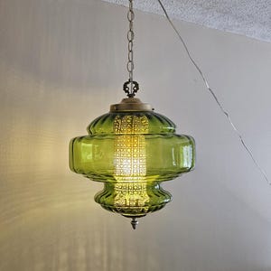 May include: A vintage green glass pendant light fixture with a brass-colored top and chain. The light has a ribbed, rounded shape and a decorative metal center. The light is hanging from the ceiling.
