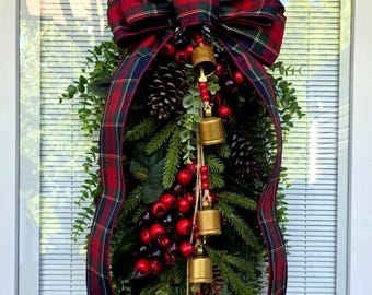 Op de afbeelding: Een kerststuk in de vorm van een swag met een grote strik van rood, groen en blauw geruit lint. Het arrangement bevat groene dennentakken, eucalyptusbladeren, bruine dennenappels, trossen rode bessen en vier hangende goudkleurige metalen bellen.