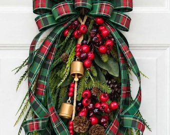 May include: A decorative door swag featuring a large bow made of green and red plaid ribbon. The arrangement includes artificial evergreen branches, clusters of bright red berries, small brown pinecones, and three hanging gold-toned bells.