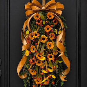 May include: A teardrop-shaped floral swag featuring orange and yellow sunflowers, small red berries, and green eucalyptus foliage. A large, wide, golden-brown velvet ribbon is tied in a bow at the top, with long, flowing tails cascading down the sides.