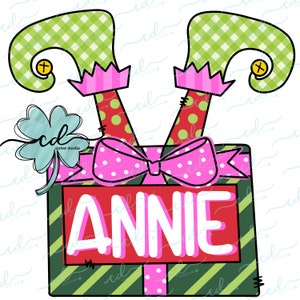 May include: A green and white striped gift box with a pink bow and two elf legs sticking out of the top. The box has the name "ANNIE" written on it in white letters.