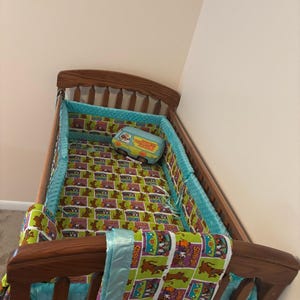 May include: A wooden baby crib with bedding featuring a Scooby-Doo theme. The bedding includes a patterned comforter, bumper pads, and a matching pillow shaped like the Mystery Machine van. The crib is made of wood and has a classic design.