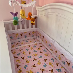 May include: A white crib with a pink and white patterned baby bedding set. The set includes a fitted sheet, bumper pads, and a mobile with hanging plush toys. The mobile has a pink canopy with a white ruffle.
