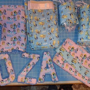 May include: A collection of handmade baby items featuring a cartoon dog print in blue, pink, and yellow. The items include blankets, rolled fabric, and letters spelling "OZA". The items are arranged on a blue cutting mat.