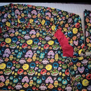 Könnte beinhalten: Ein schwarzes Stoffset mit einem farbenfrohen Muster von Sesame Street-Charakteren. Das Set enthält ein Spannbetttuch, ein Bettlaken und einen Kissenbezug. Das Spannbetttuch hat rote Akzente. Das Muster zeigt Charaktere wie Krümelmonster, Elmo und Big Bird.