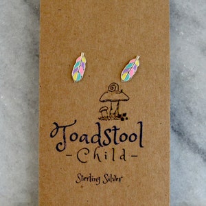 May include: A pair of sterling silver earrings featuring a colorful feather design. The earrings are on a brown card with the text "Toadstool Child - Sterling Silver".