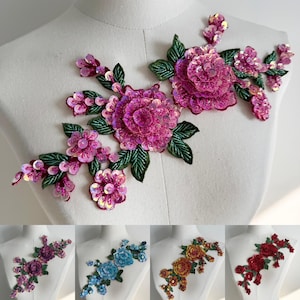 Può includere: Applicazione floreale con paillettes rosa e foglie verdi. L'applicazione presenta grandi fiori dettagliati e gruppi floreali più piccoli. Ulteriori varianti di colore sono mostrate di seguito, tra cui viola, blu, oro e rosso.