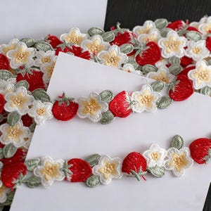 May include: Embroidered trim with a repeating pattern of red strawberries, white and yellow flowers, and green leaves. The trim is likely made of fabric and is suitable for sewing or crafting projects. The design is detailed and colourful.