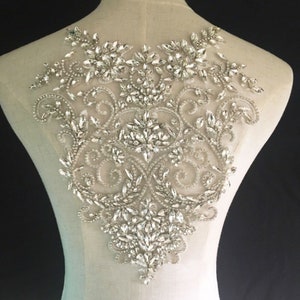May include: A large, intricate, silver-toned rhinestone applique with a floral design. The applique is made of small, clear rhinestones and silver beads, and it is perfect for adding a touch of sparkle to any garment.