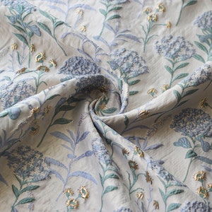 May include: A white fabric with a blue and green floral pattern. The fabric has a textured, embroidered look.