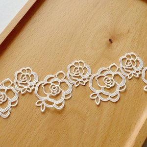Può includere: Bordino in pizzo bianco con un delicato disegno floreale che presenta un motivo ripetitivo di piccole rose. Il pizzo è su una superficie di legno.