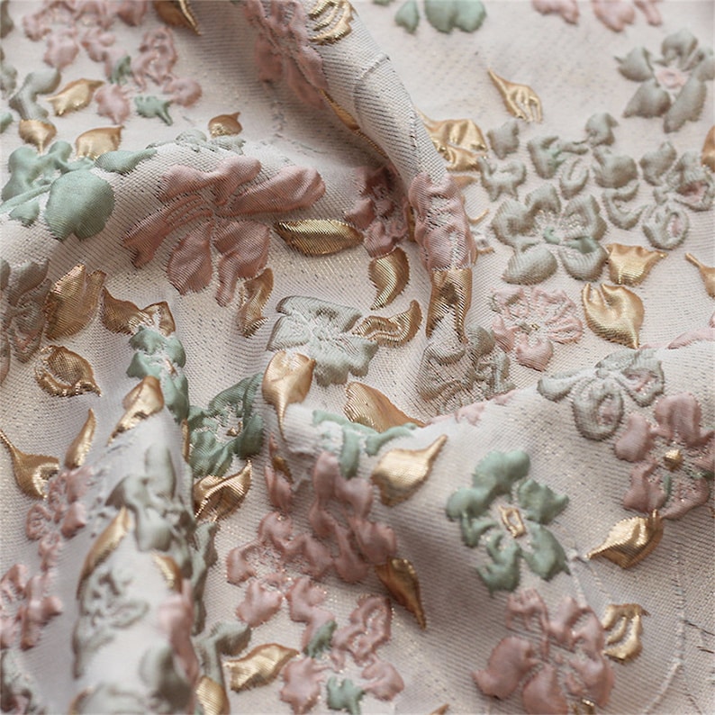 May include: Close-up of a cream-colored fabric with a raised floral pattern. The flowers are pink, green, and gold.