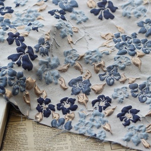 May include: A close-up of a white fabric with a blue and gold floral pattern. The fabric is textured and has a raised design.