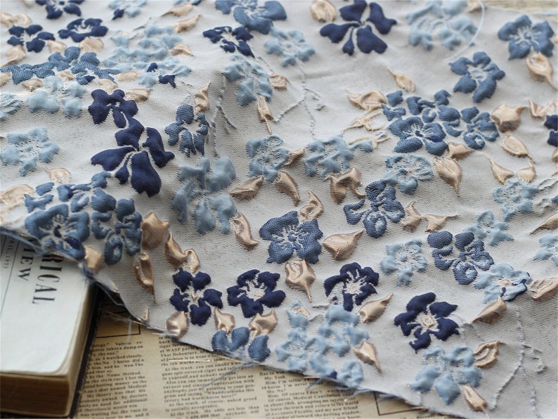 May include: A close-up of a white fabric with a blue and gold floral pattern. The fabric is textured and has a raised design.