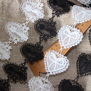 May include: A close-up of black and white lace trim with a heart design. The trim is shown on a beige fabric background.