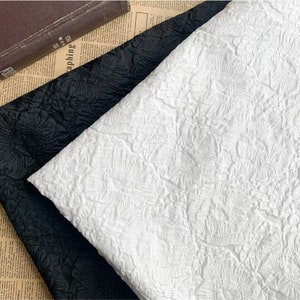 May include: Close-up of two fabric swatches, one black and one white, with a raised floral pattern. The fabrics are folded and layered, showcasing the texture and design. A vintage book rests in the background.