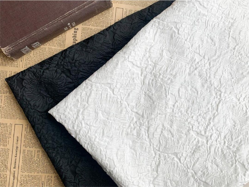 May include: Close-up of two fabric swatches, one black and one white, with a raised floral pattern. The fabrics are folded and layered, showcasing the texture and design. A vintage book rests in the background.