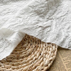 May include: Close-up of white fabric with a textured, floral pattern. The fabric is draped over a woven basket. The fabric has a soft, ruffled appearance. The basket is made of natural fibers.