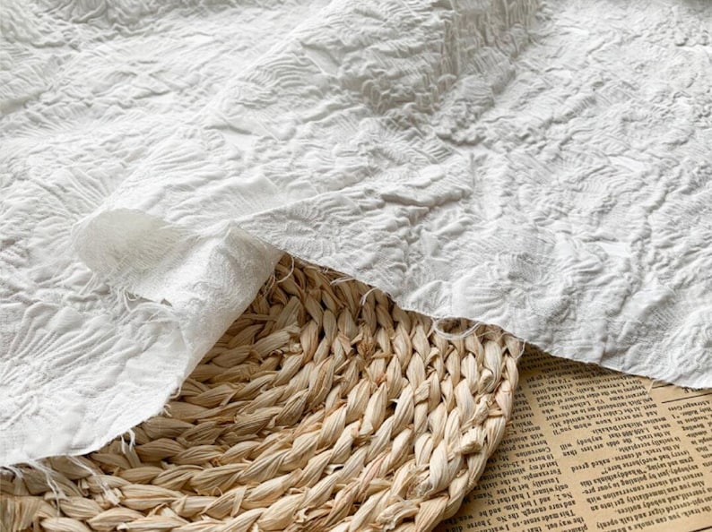 May include: Close-up of white fabric with a textured, floral pattern. The fabric is draped over a woven basket. The fabric has a soft, ruffled appearance. The basket is made of natural fibers.