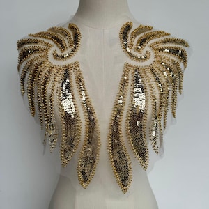 May include: Gold sequin and bead applique with a wing-like design. The applique has a sheer, white mesh backing and is heavily embellished with gold sequins and small, gold beads. The design is symmetrical, with each side mirroring the other.