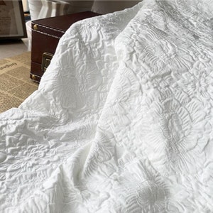 May include: White fabric with a raised floral pattern. The fabric is draped, showing the texture and design. The material appears to be thick and possibly used for upholstery or bedding.