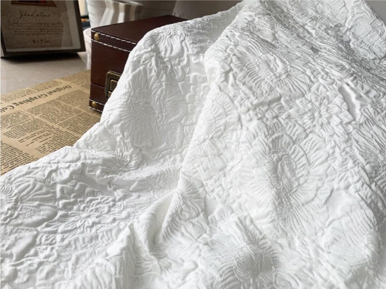 May include: White fabric with a raised floral pattern. The fabric is draped, showing the texture and design. The material appears to be thick and possibly used for upholstery or bedding.