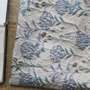 May include: White fabric with a repeating pattern of blue and green embroidered flowers. The flowers are stylized and have a 3D effect. The fabric has a subtle texture.