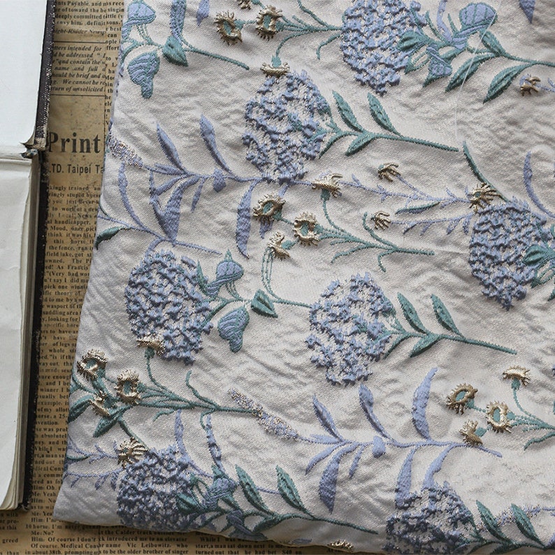 May include: White fabric with a repeating pattern of blue and green embroidered flowers. The flowers are stylized and have a 3D effect. The fabric has a subtle texture.