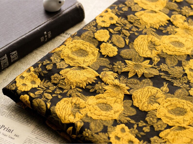 May include: Black fabric with a gold floral pattern. The fabric has a raised texture, giving it a 3D effect.