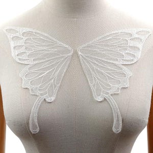 Könnte beinhalten: Weiße Spitzen-Schmetterlingsapplikation zum Nähen oder Basteln. Der Schmetterling ist zart und filigran mit einem detaillierten Flügelmuster.