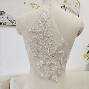 May include: A white beaded floral applique with silver accents on a sheer fabric background. The applique features a delicate branch with leaves and small beads.