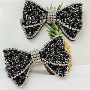 May include: Two black bow embellishments, heavily embellished with small, sparkling black and silver rhinestones. The bows have a layered design and are accented with a silver rhinestone center. The bows are likely for hair accessories or fashion.